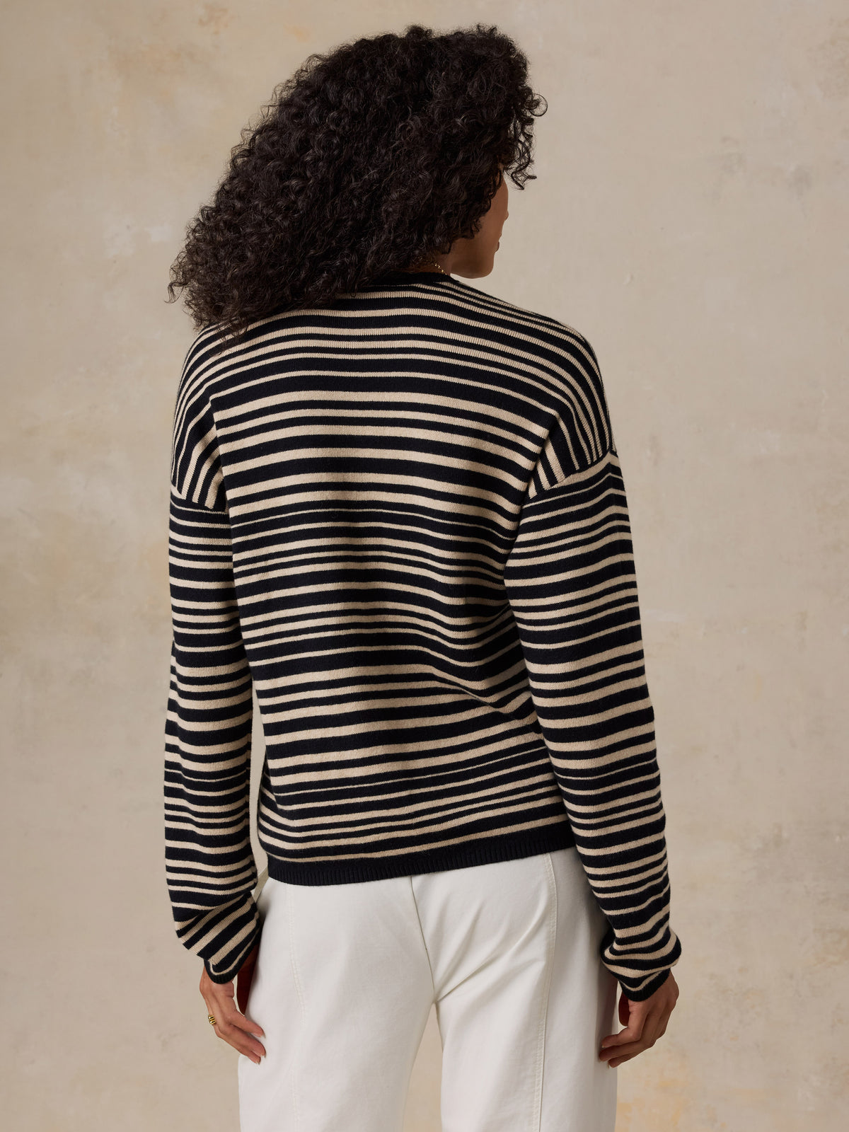 A woman shown from the back wearing a black and cream horizontal striped knit sweater and white trousers, standing against a textured beige wall., image 4 of 6