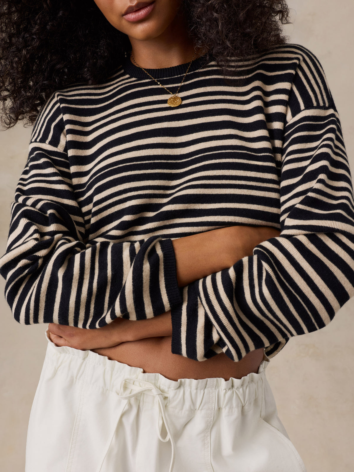 A close-up of a woman wearing a cropped long-sleeve knit sweater with horizontal black and cream stripes, paired with white drawstring pants and a small gold pendant necklace., image 2 of 6