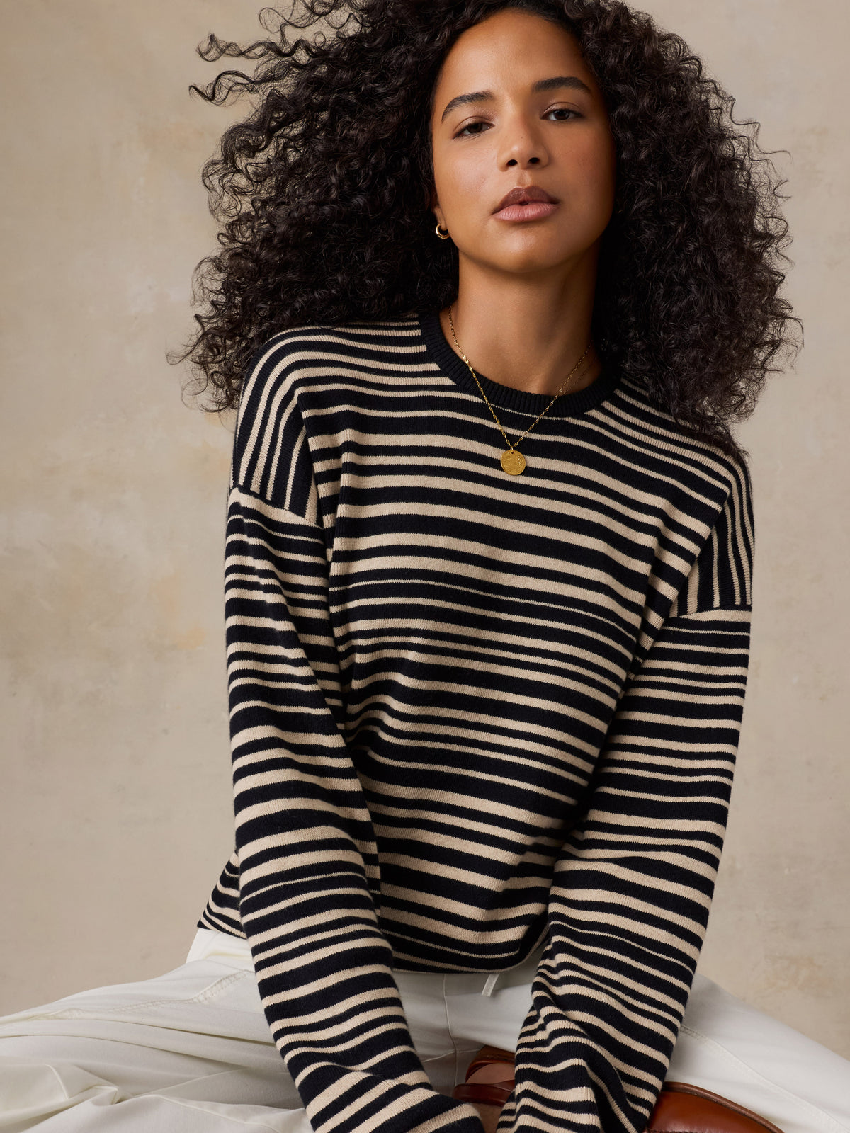 A woman with curly dark hair wearing a black and cream horizontal striped long-sleeve knit sweater, paired with white pants, posed against a textured beige wall., image 5 of 6