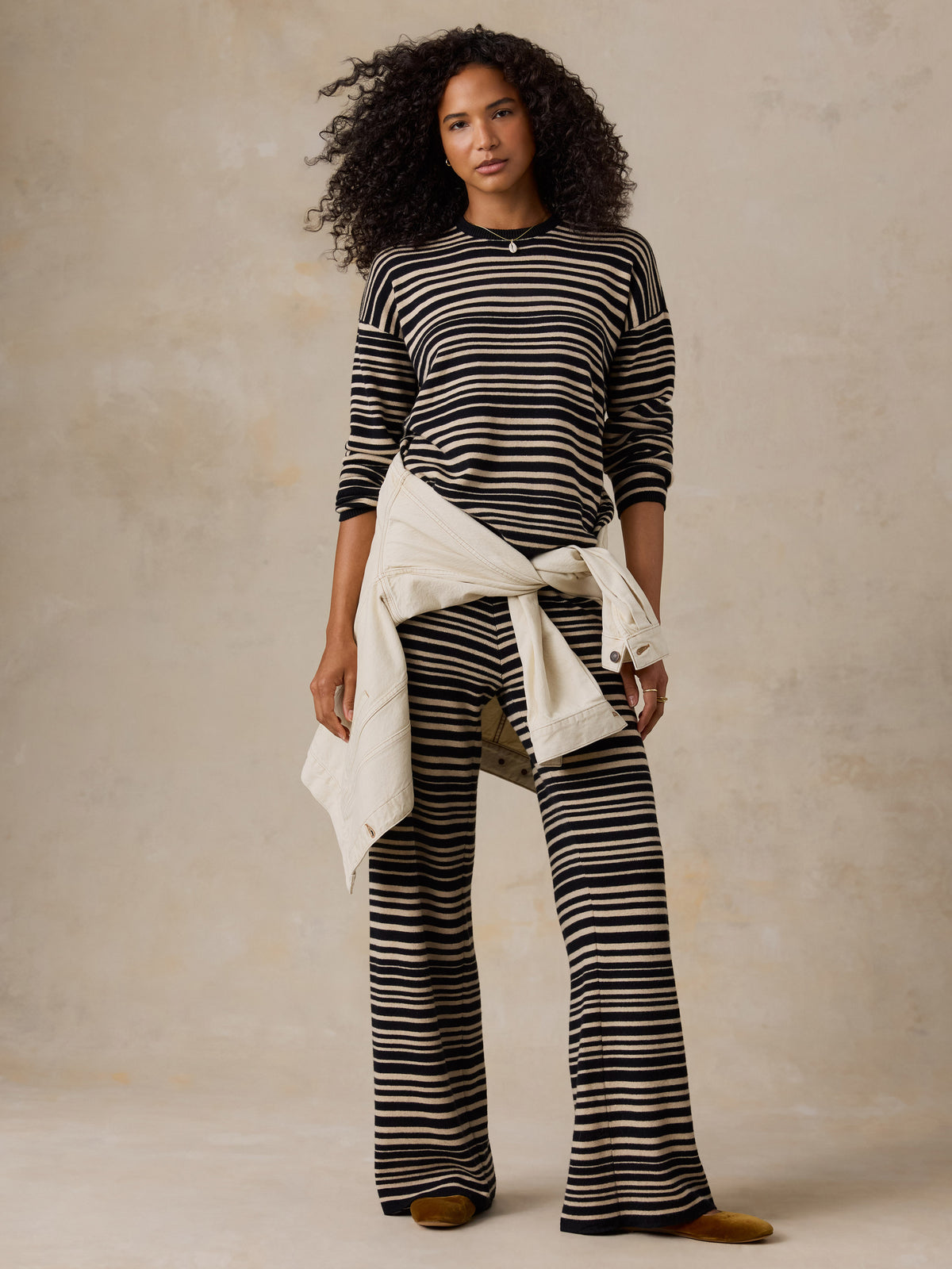 A model with curly black hair stands against a textured beige studio backdrop, wearing a black and cream horizontal striped long-sleeve top and matching striped wide-leg pants, with a cream shirt tied around her waist., image 6 of 6
