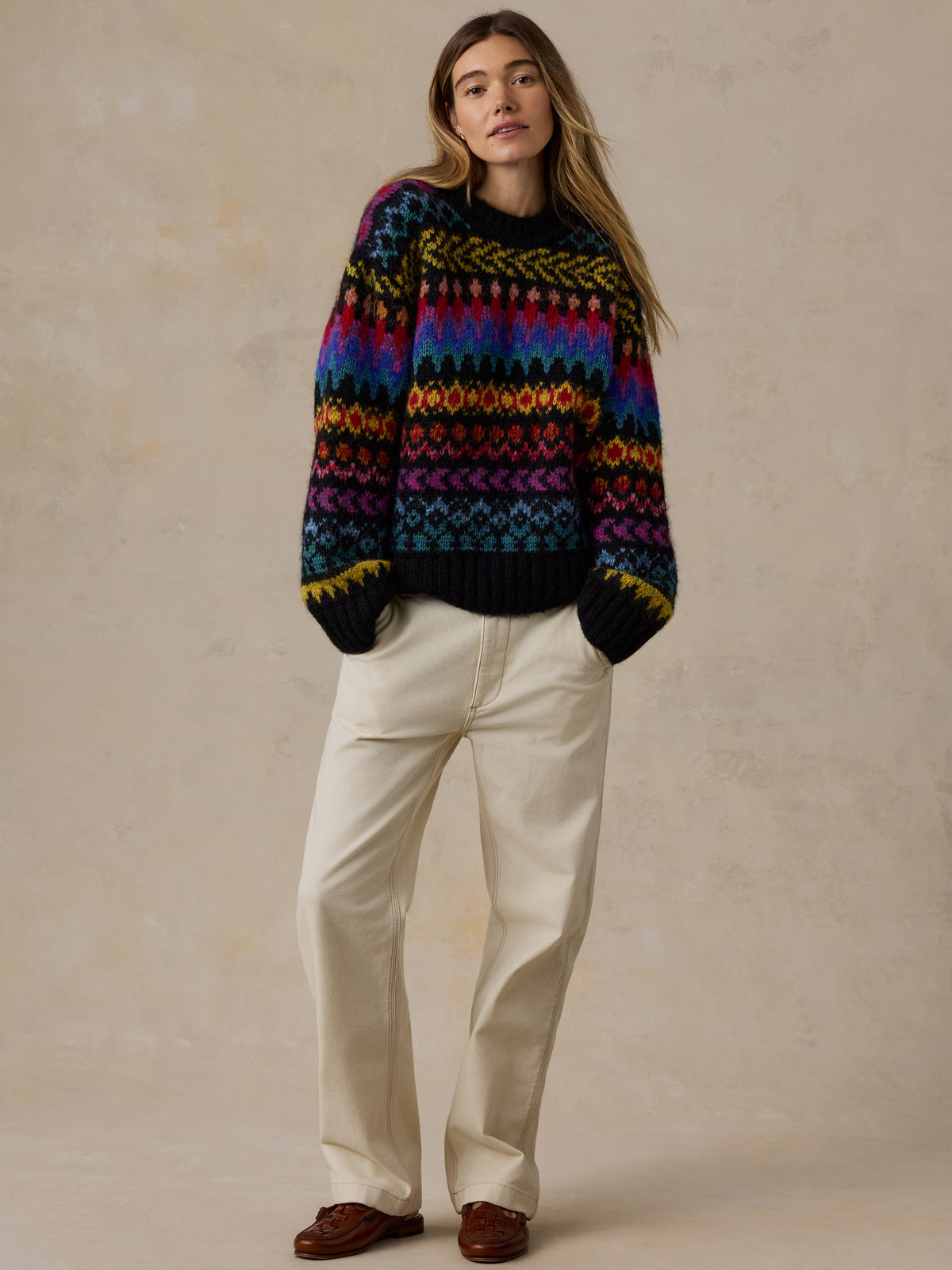 A woman stands against a textured beige studio backdrop wearing a colorful Fair Isle knit sweater with a black base and rainbow stripes, paired with cream trousers and brown shoes., image 7 of 7