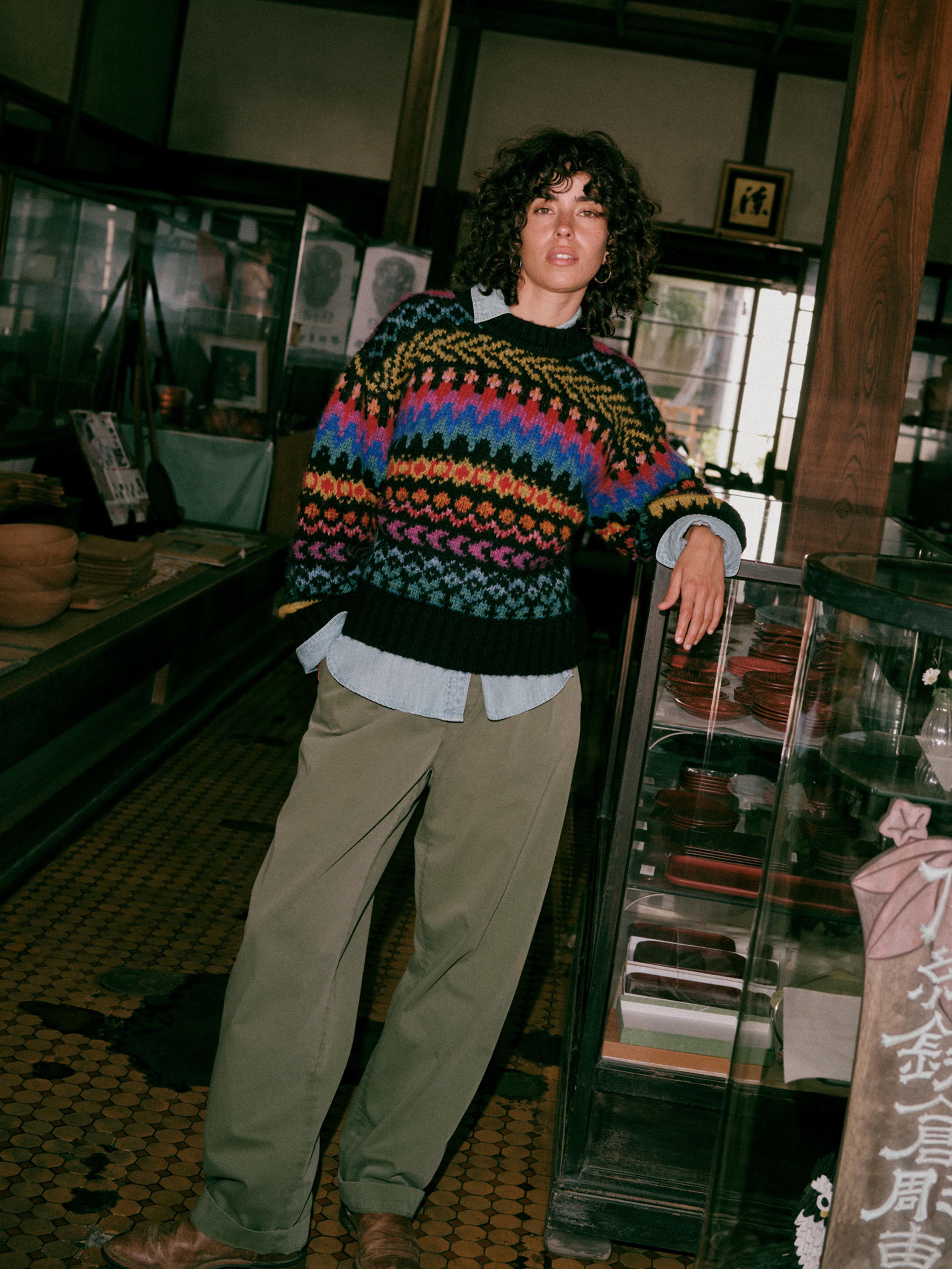 A woman standing in a shop interior wearing a colorful patterned knit sweater over a light blue shirt, with loose olive pants and brown shoes.