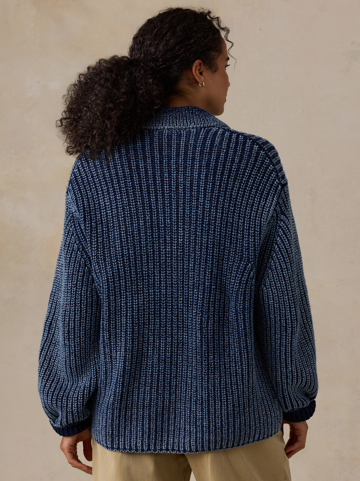 A woman shown from the back wearing a dark blue ribbed knit cardigan or sweater with beige pants, standing against a plain beige wall., image 5 of 8