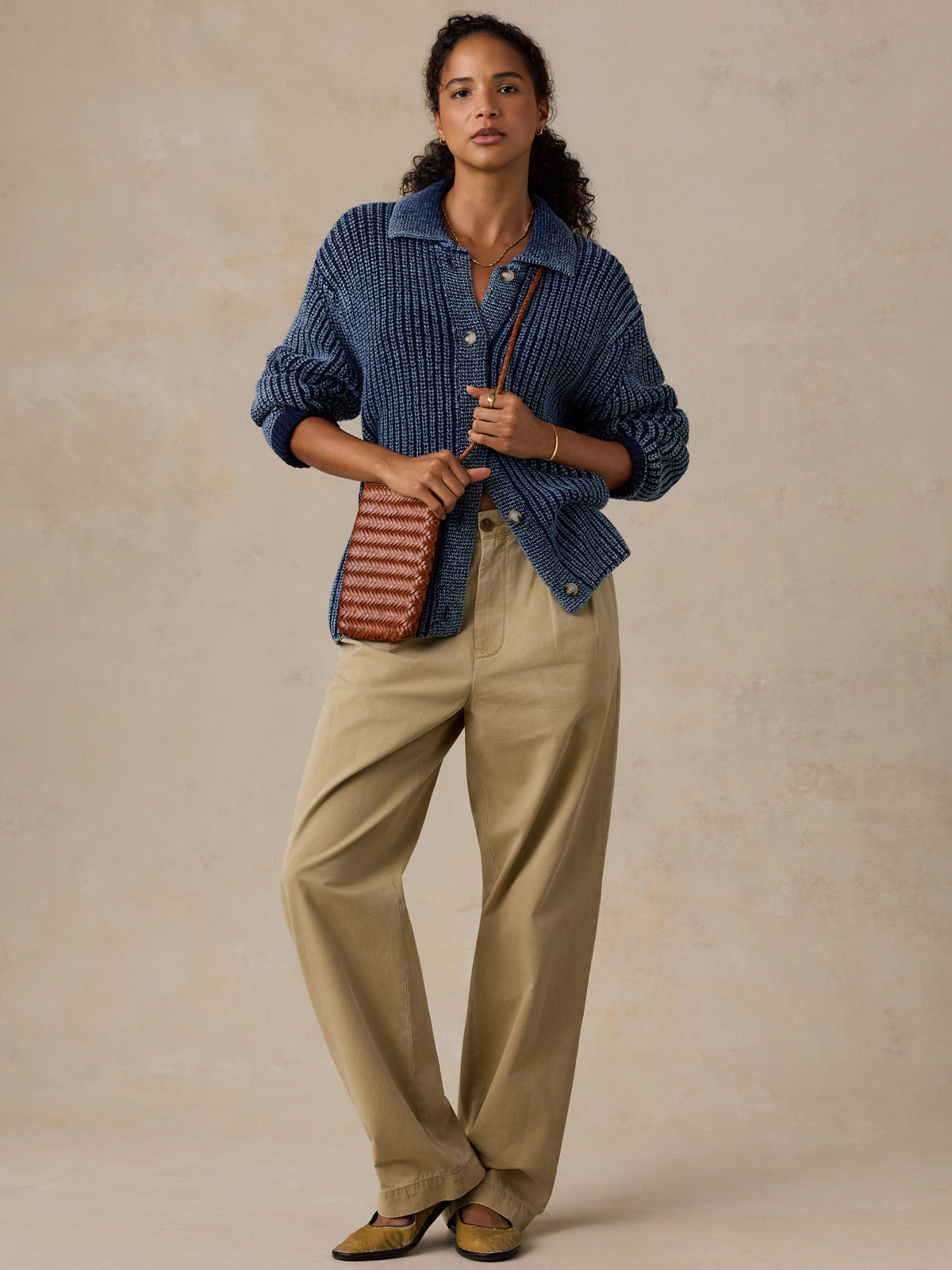 A woman stands against a textured beige studio backdrop wearing a blue ribbed knit button-up shirt, beige wide-leg pants, and tan loafers, while holding a brown quilted shoulder bag., image 3 of 8