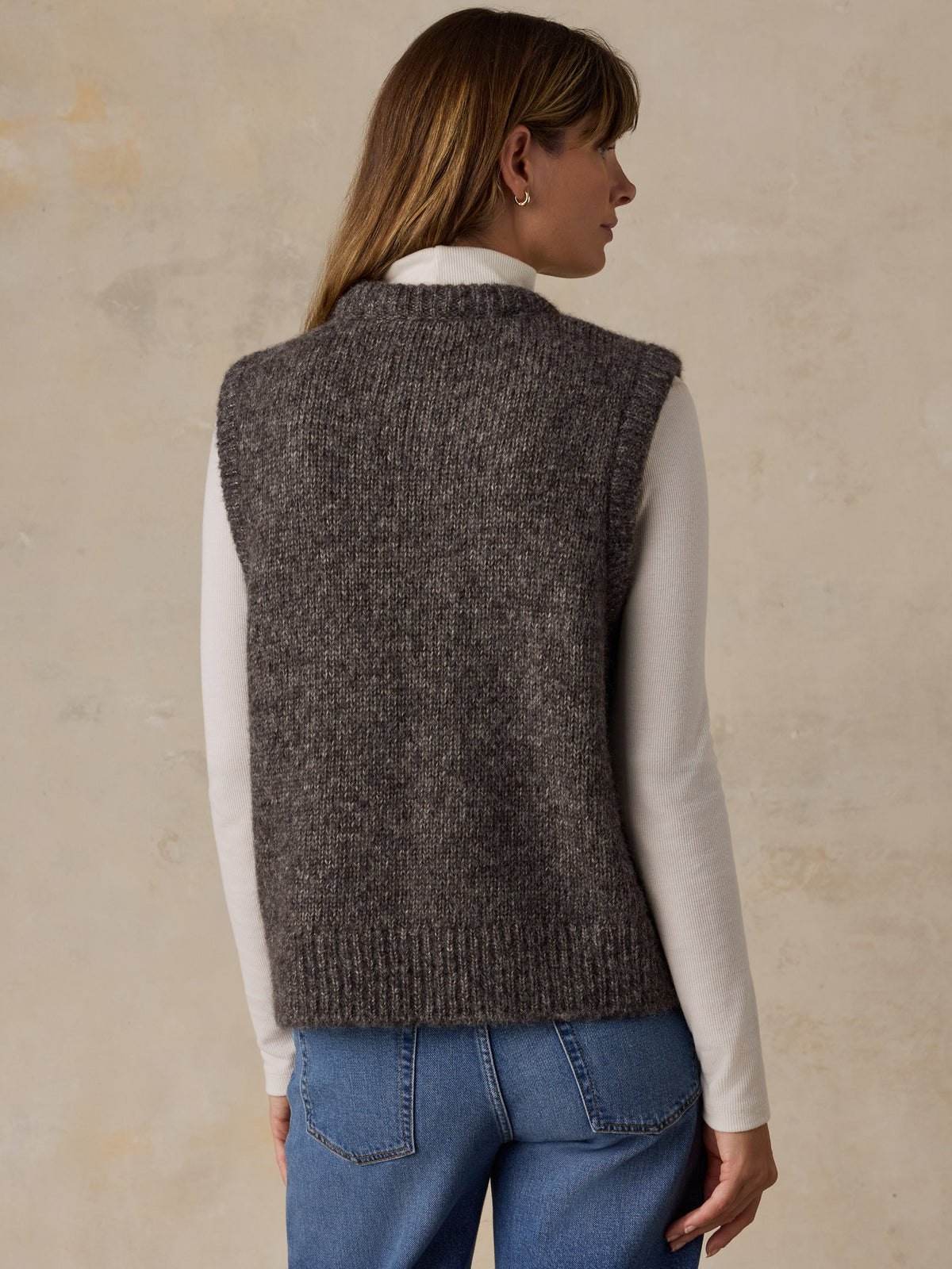 A woman viewed from behind wearing a grey sweater vest with a white turtleneck and blue jeans, image 3 of 5