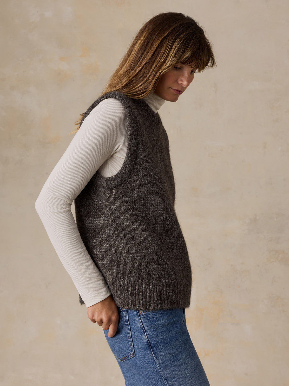 A woman wearing a grey sweater vest with a white turtleneck and blue jeans, image 4 of 5