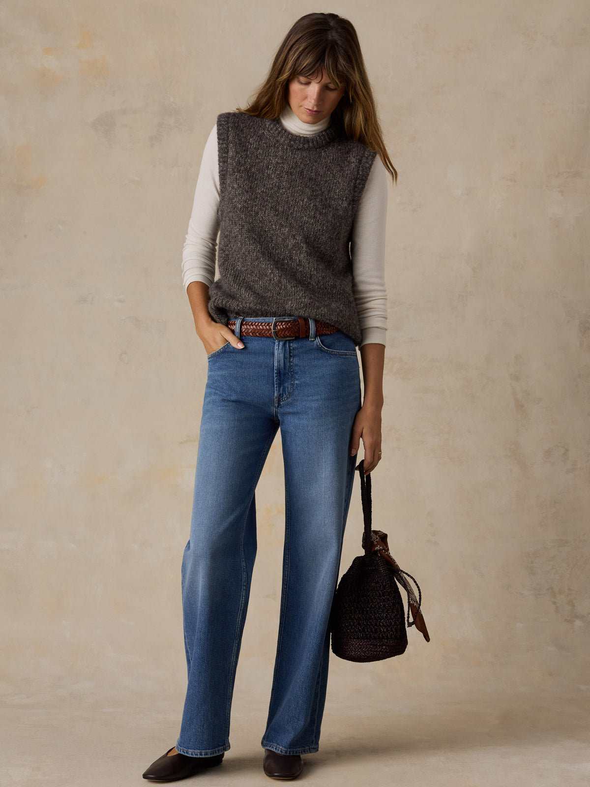 A woman wearing a grey sweater vest with a white turtleneck, blue jeans, and flats, image 2 of 5