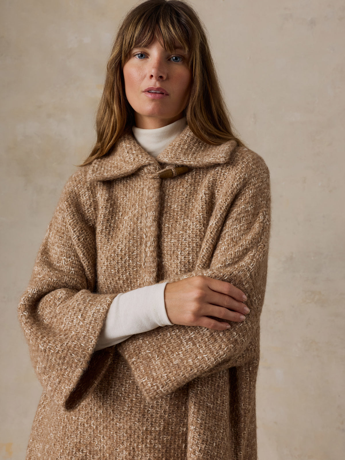 A woman wearing a beige sweater poncho over a white turtleneck, image 6 of 7