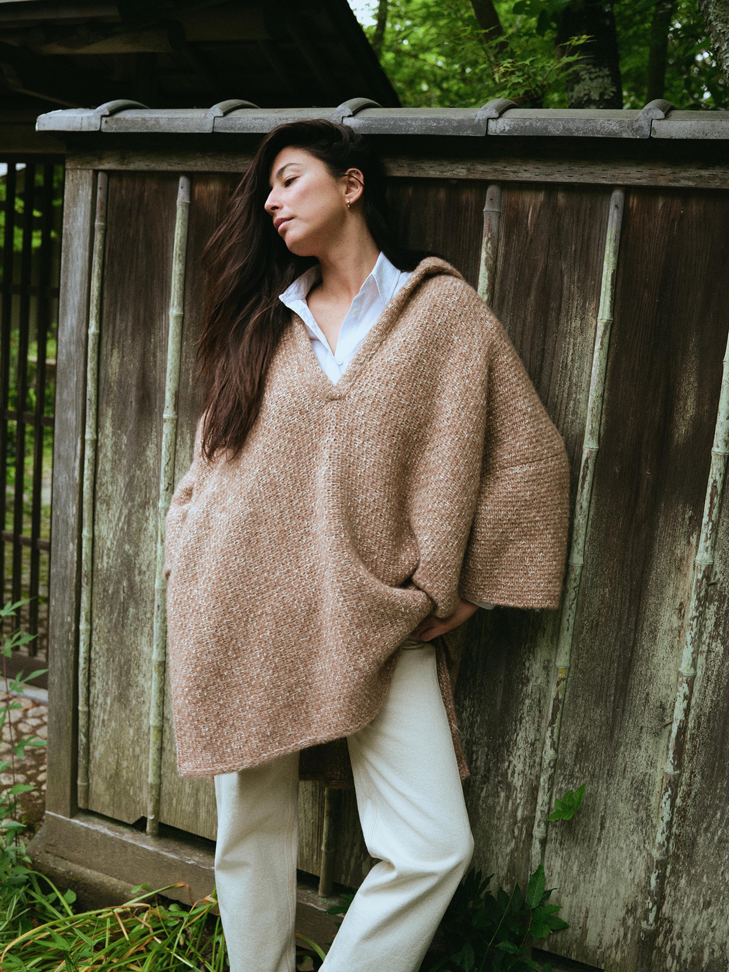 A woman wearing a chunky knit beige hooded poncho over a white collared shirt and cream pants, leaning against a weathered wooden fence outdoors.