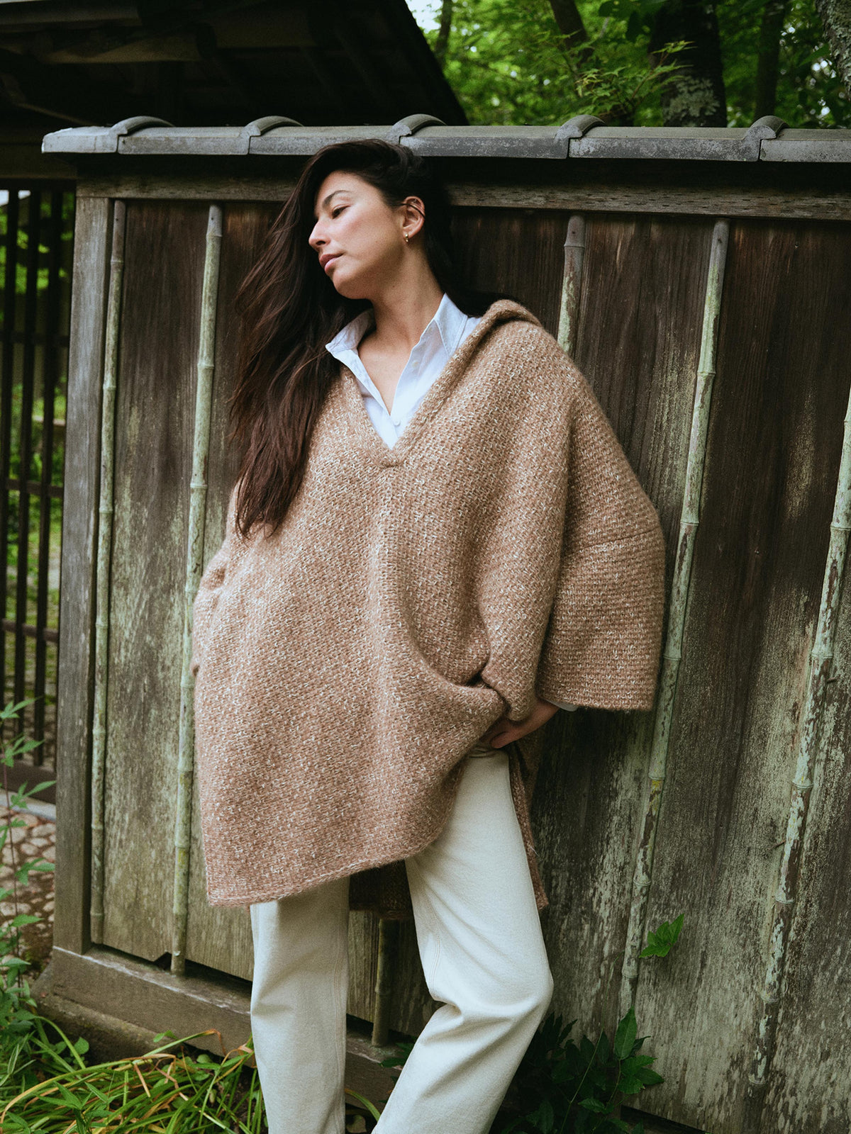 A woman wearing a chunky knit beige hooded poncho over a white collared shirt and cream pants, leaning against a weathered wooden fence outdoors., image 1 of 7