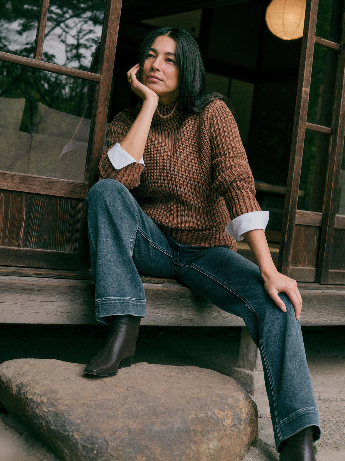 A woman sits on a stone step in front of a wooden building, wearing a brown ribbed knit sweater over a white collared shirt, blue jeans, and dark ankle boots., image 1 of 7
