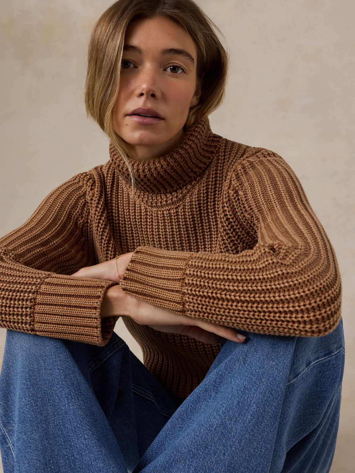 Woman wearing a brown knit turtleneck sweater and blue jeans against a beige studio background, image 7 of 7