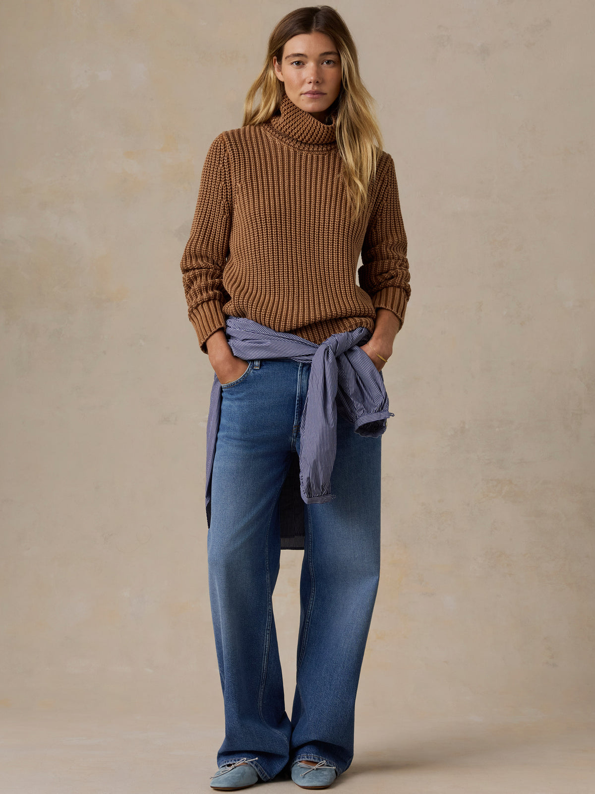 Woman wearing a brown knit turtleneck sweater and blue jeans against a beige studio background, image 5 of 7