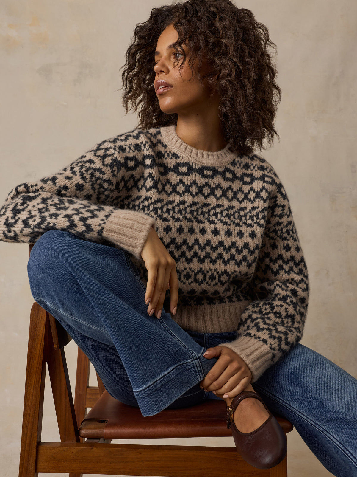 A woman sitting on a chair wearing a beige printed sweater, blue jeans, and flats, image 4 of 7