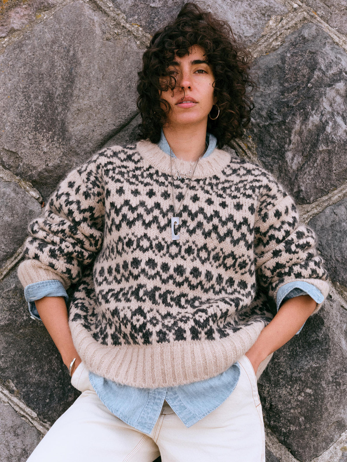 A model with curly dark hair wearing a beige and black patterned knit sweater over a light blue shirt, paired with white pants, leaning against a gray stone wall., image 1 of 7