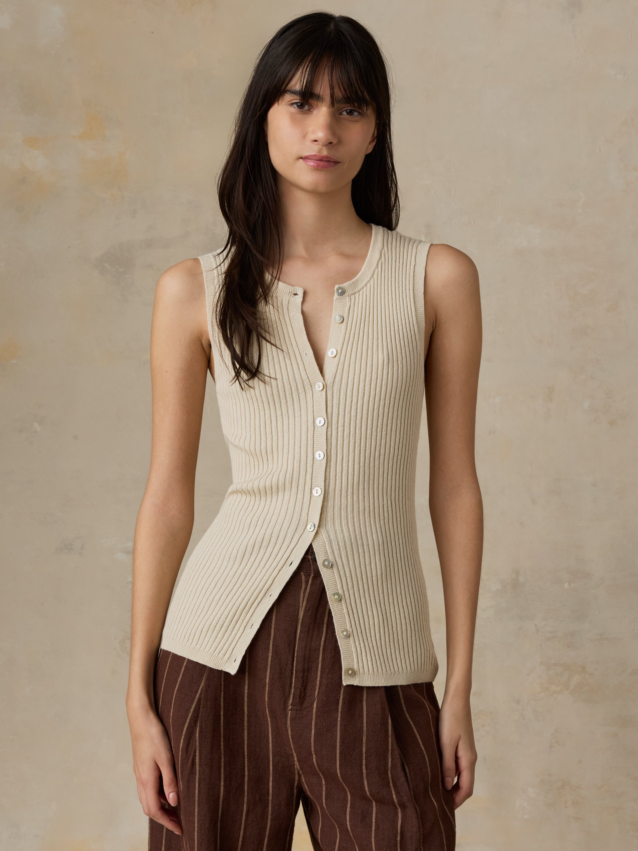 Woman wearing a beige sleeveless button-up top with brown pants against a beige background