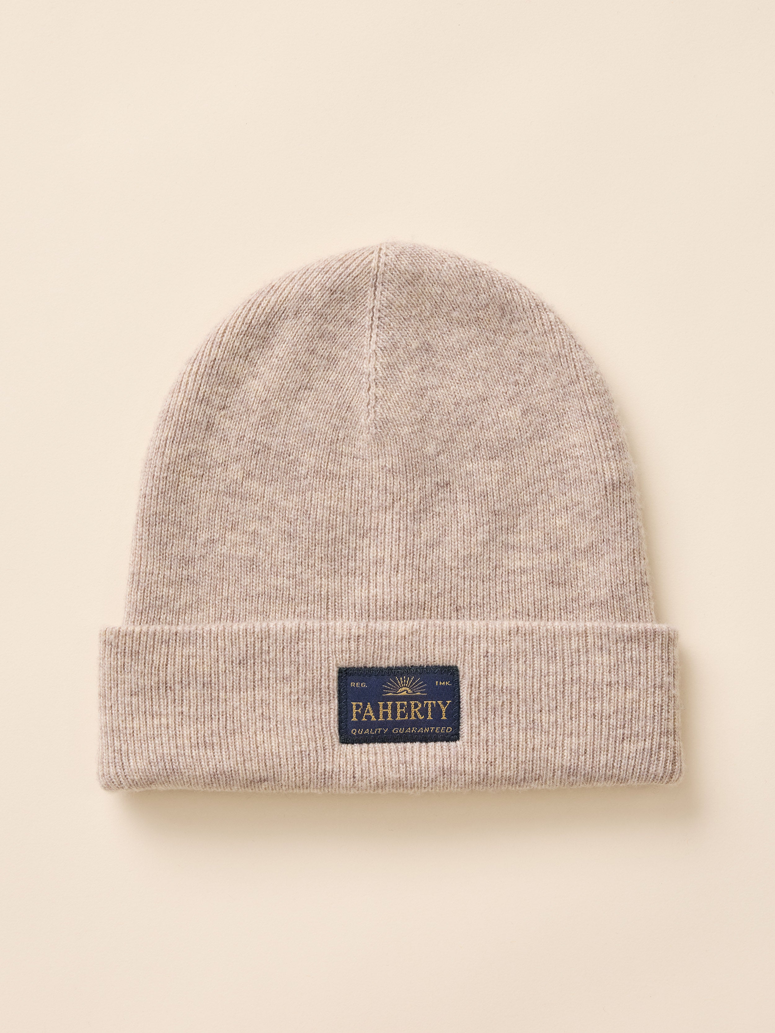 A light beige knit beanie hat with a folded cuff, featuring a small navy blue brand patch that reads 'FAHERTY'. The hat is photographed flat against a solid, warm cream background.