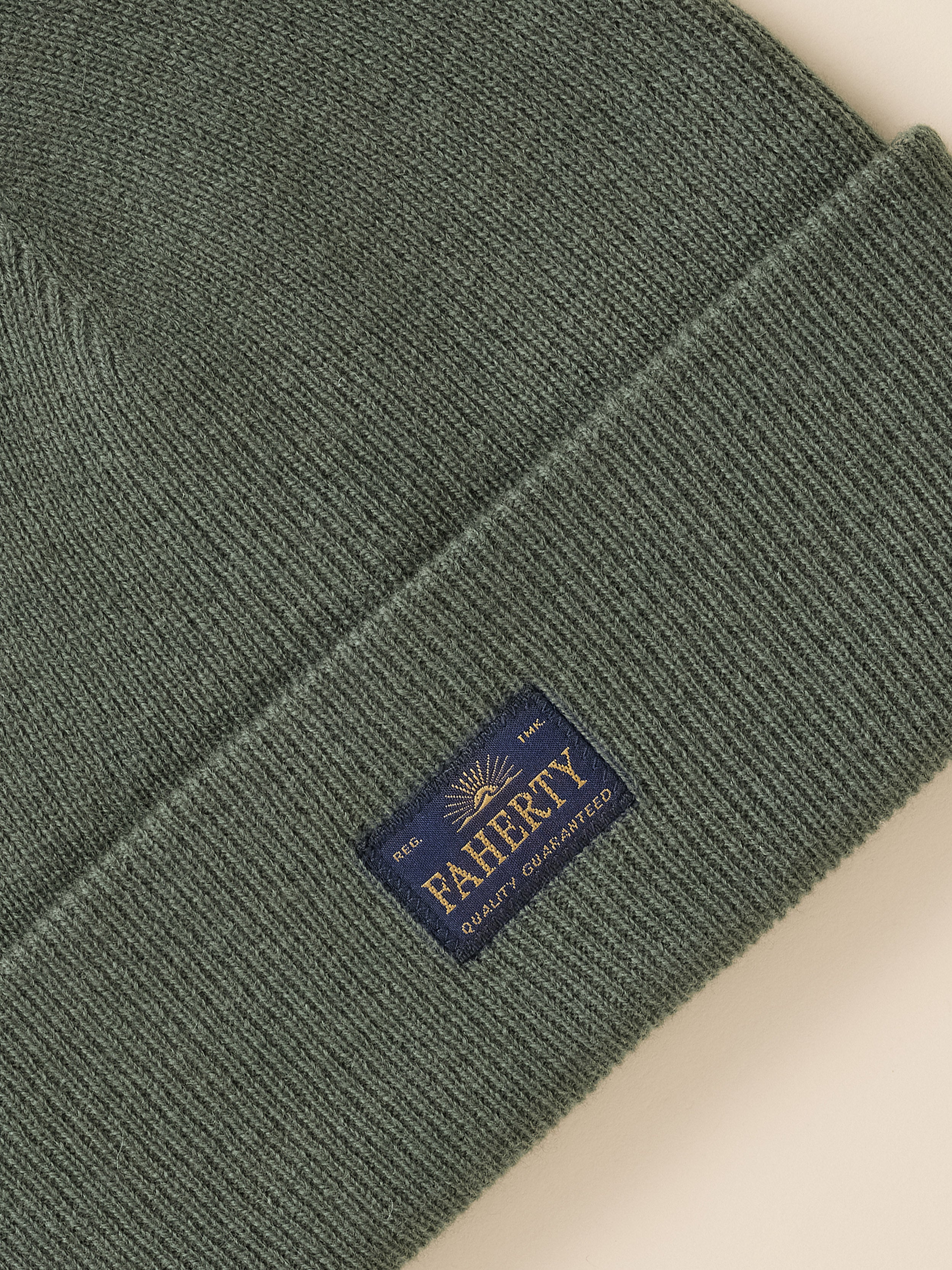Close-up of a ribbed knit beanie in dark olive green, featuring a navy blue sewn-on brand patch that reads 'PAHRTY QUALITY GUARANTEED' with a gold sun logo, set against a light beige background.