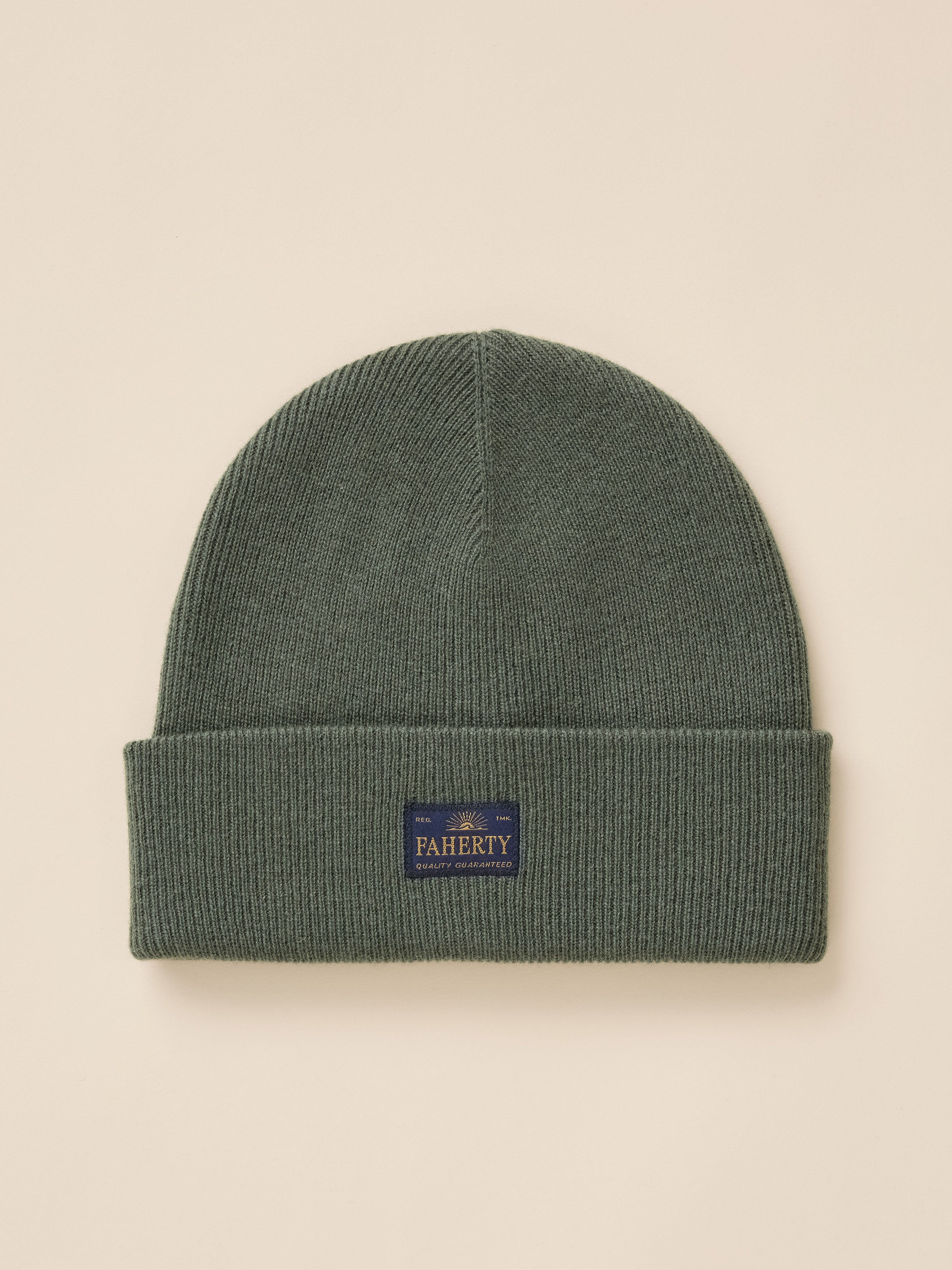 A dark green ribbed knit beanie hat with a folded cuff, featuring a small navy blue FAHERTY brand patch on the front, set against a solid beige background.