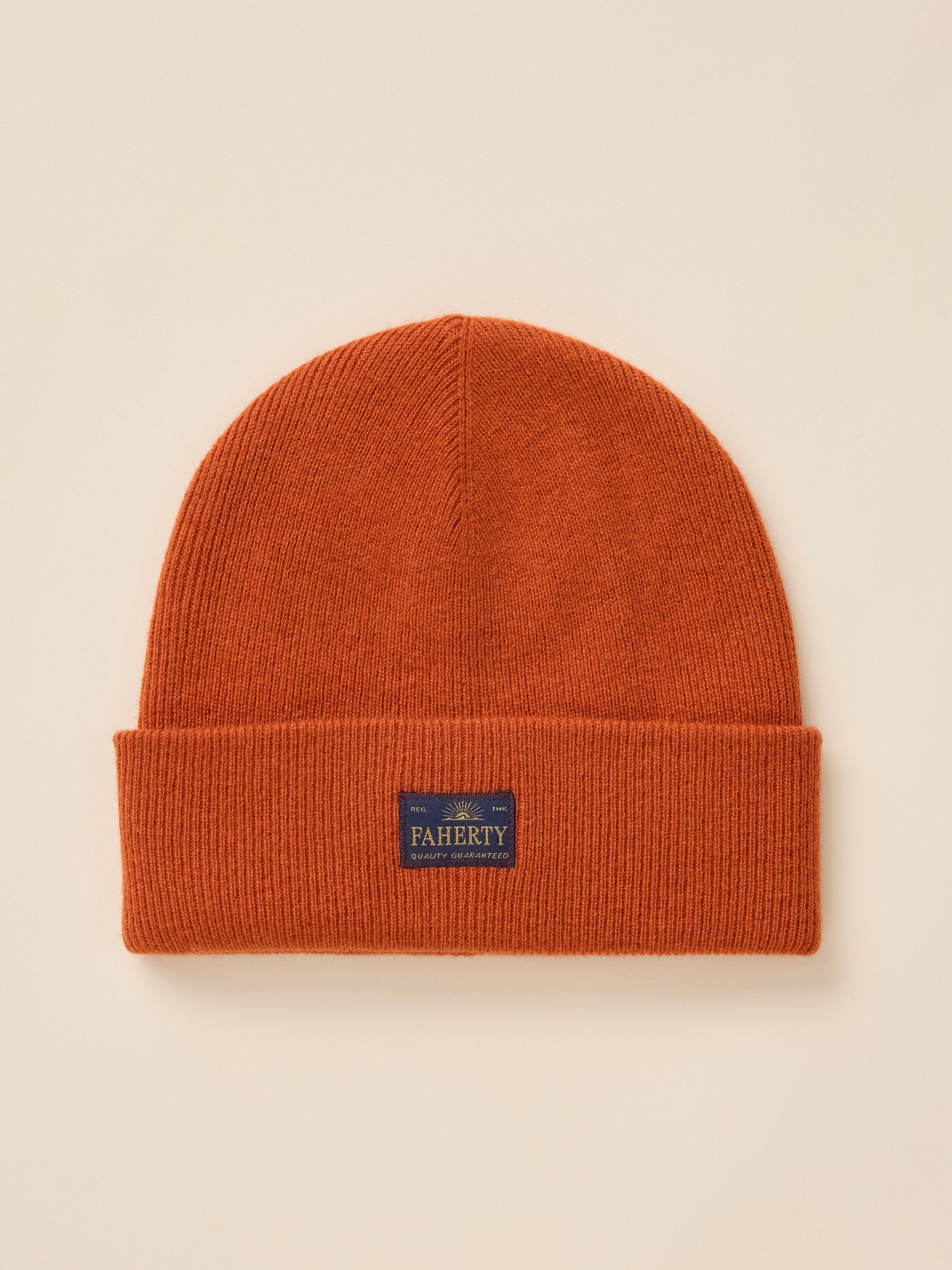 An orange ribbed knit beanie hat with a folded cuff, featuring a small navy blue FAHERTY brand patch on the front, set against a solid beige background.