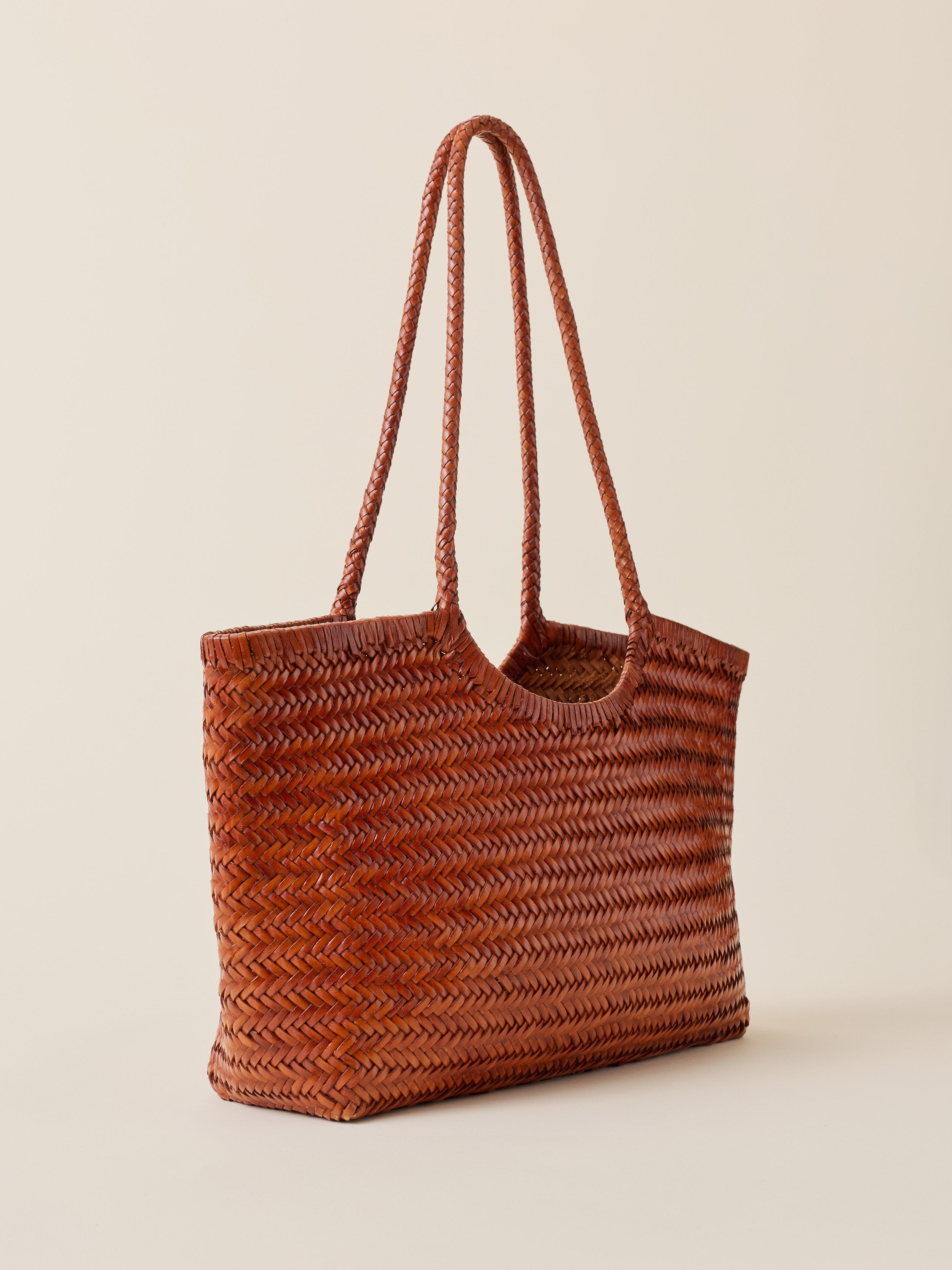 A brown woven straw tote bag with two braided shoulder straps, shown on a solid light beige background.