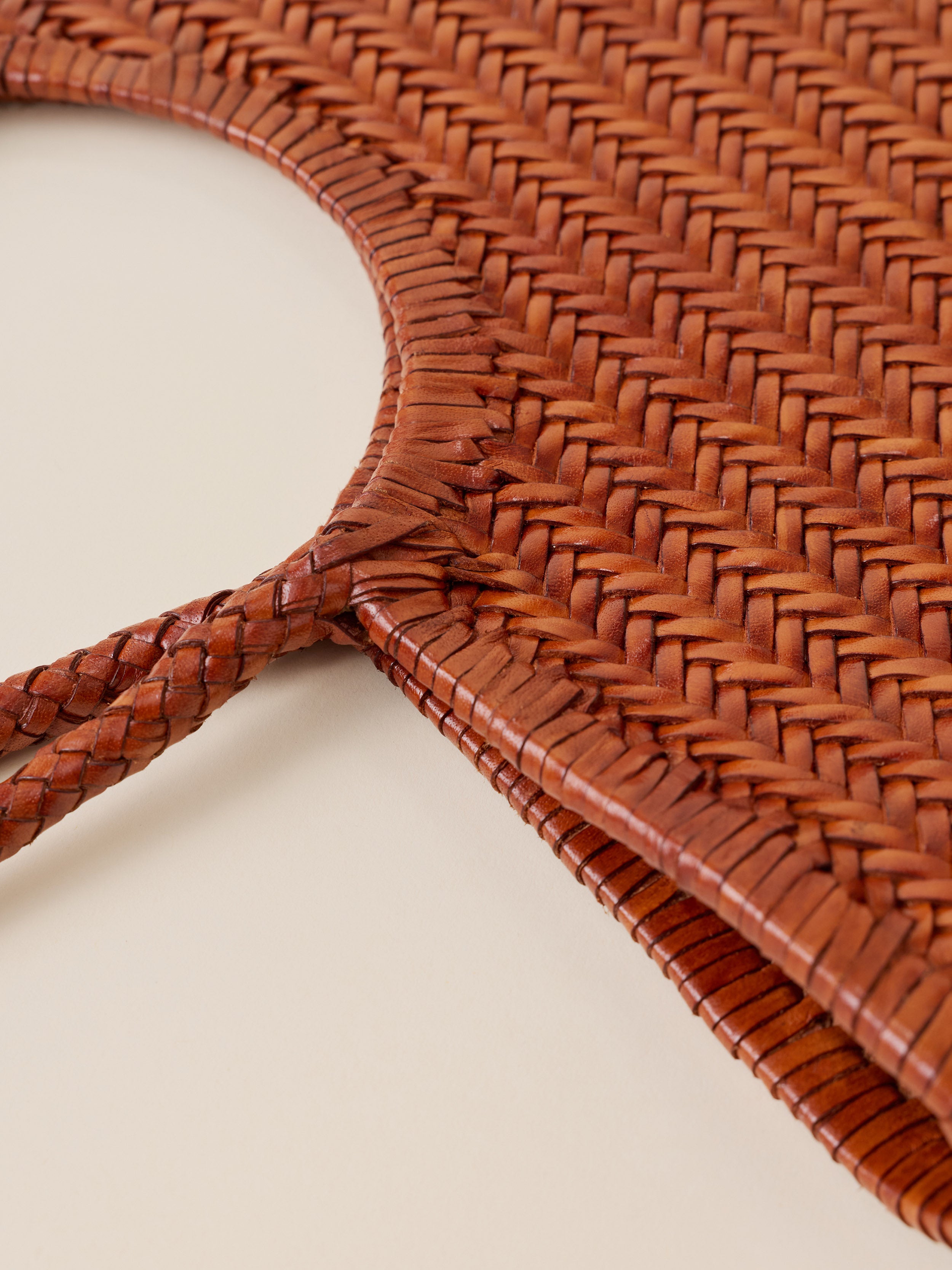 Close-up of a brown woven leather accessory, showing the detailed braided texture and curved edge on a light beige background.