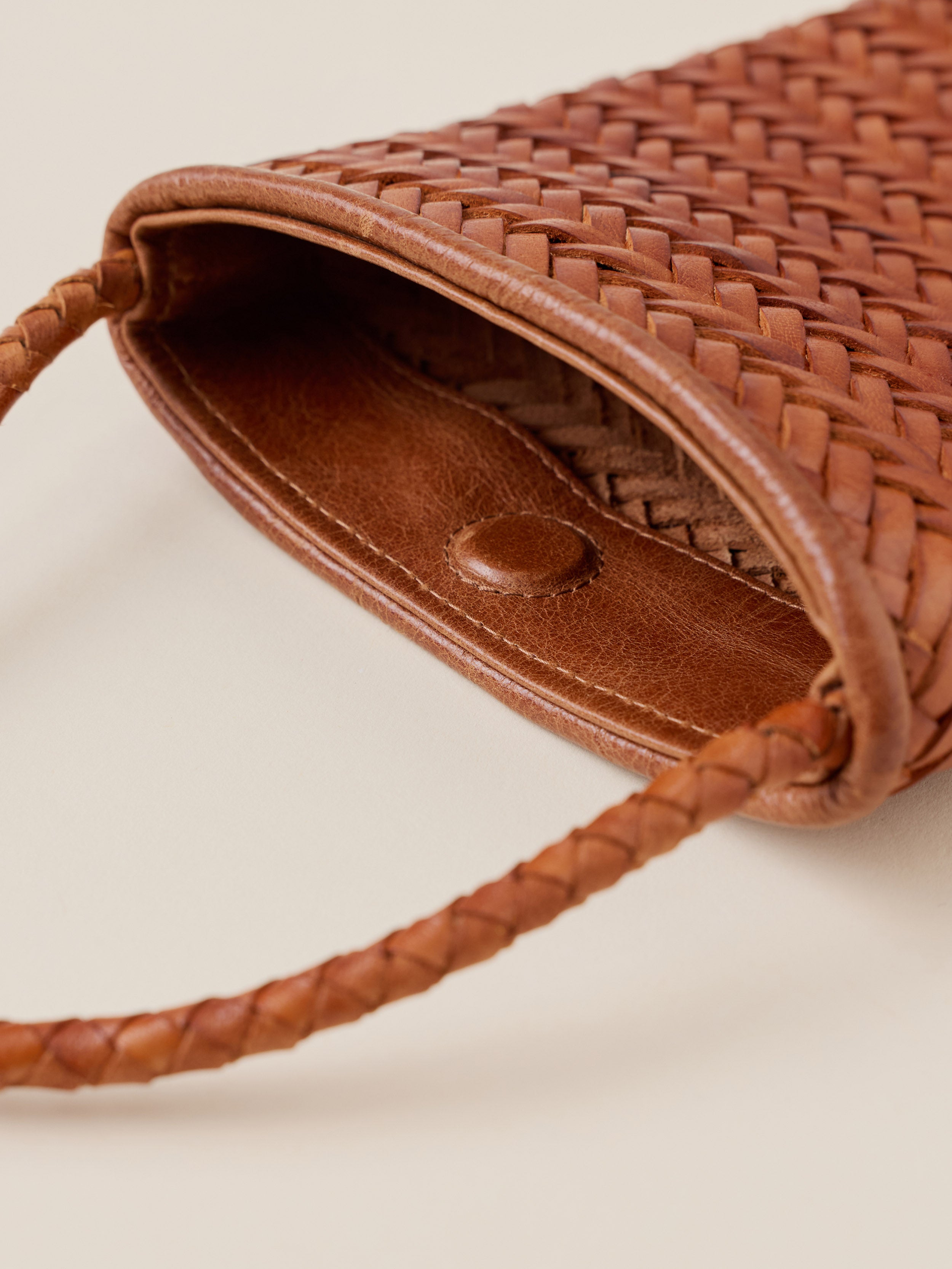 Close-up of a brown woven leather bag with a braided shoulder strap and a smooth leather interior, photographed on a light beige background.