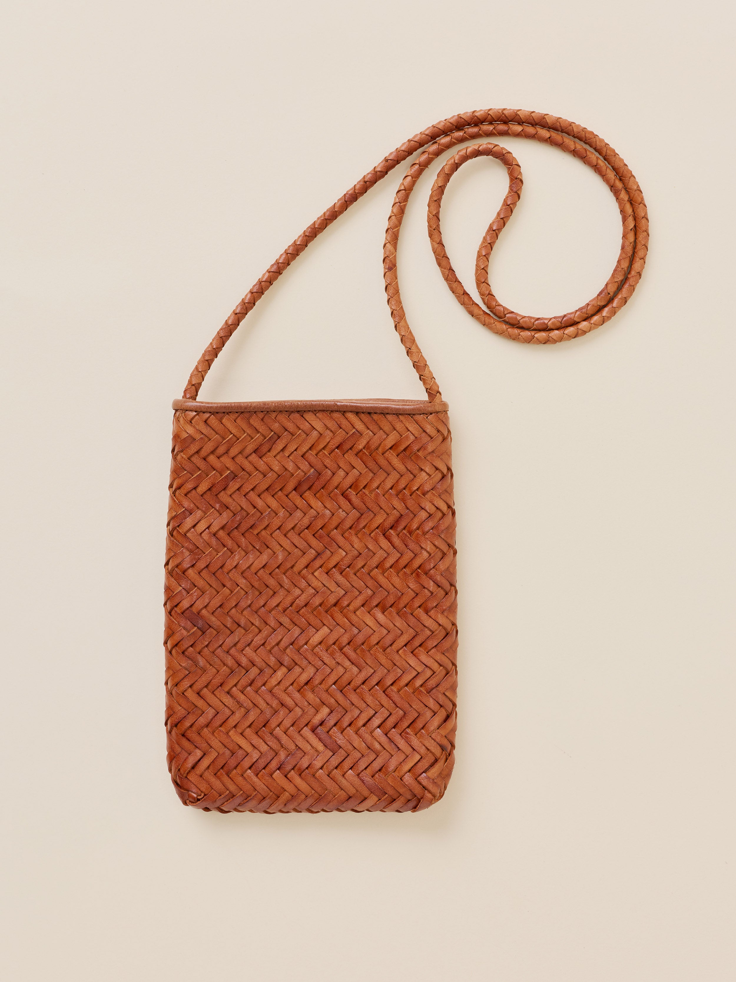 A small rectangular woven leather shoulder bag in a warm cognac brown, featuring a herringbone pattern and a long braided strap, set against a solid light beige background.