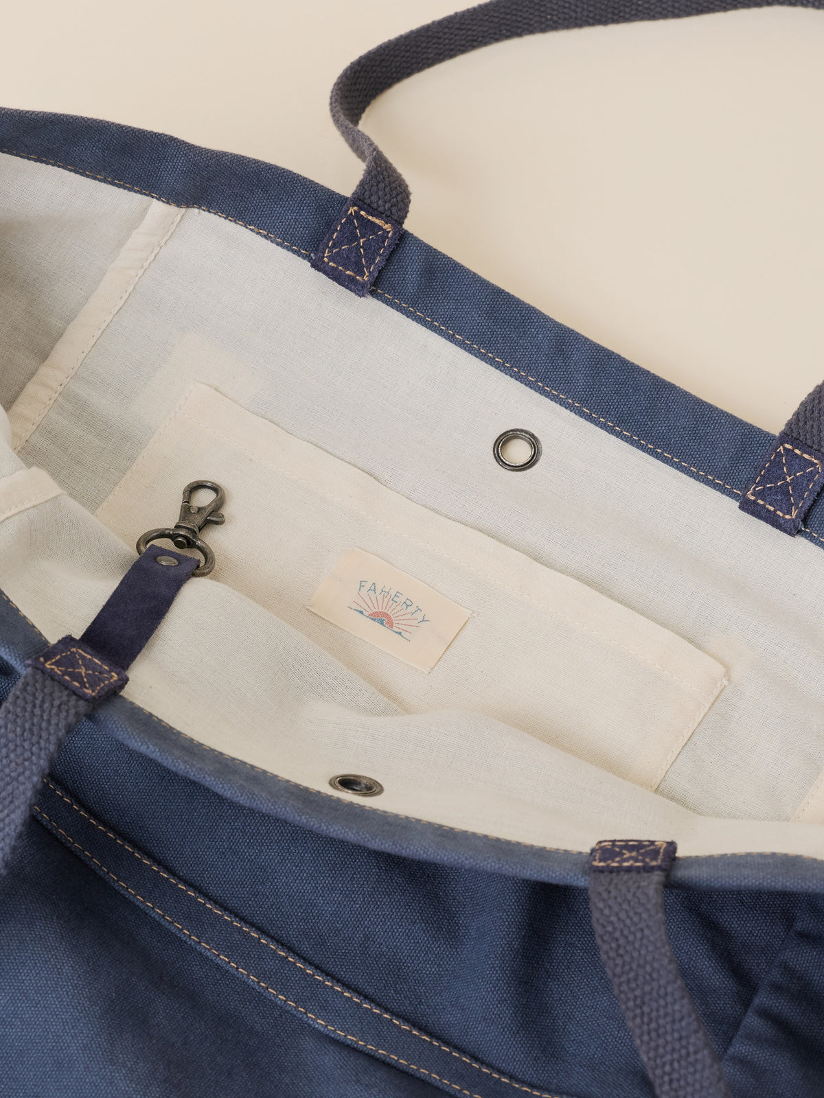Close-up of a navy blue canvas tote bag showing the interior with a cream-colored fabric pocket, metal grommets, and a small sewn-in Faherty brand label.