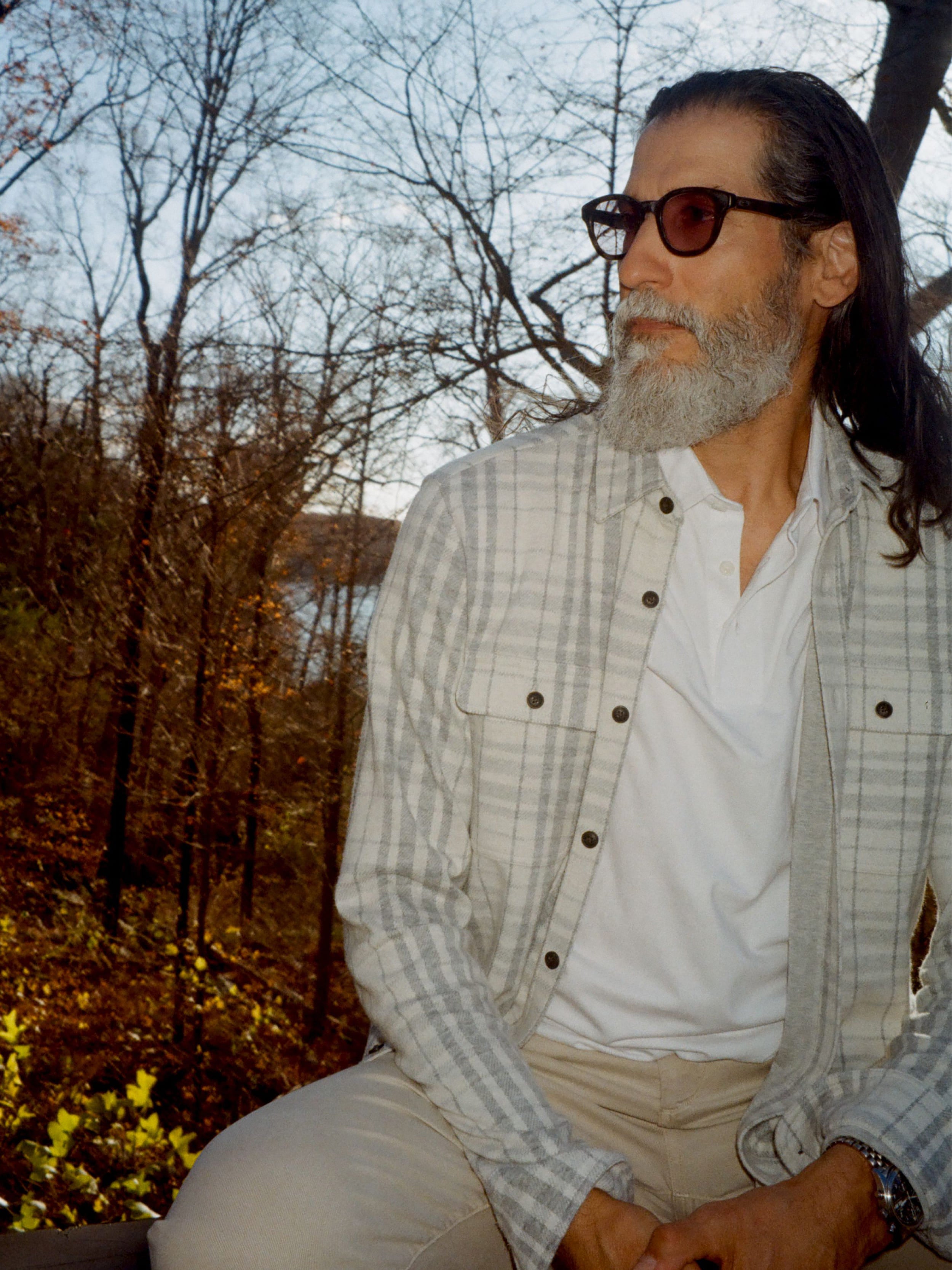 Man wearing a white checkered long sleeve button up shirt and sunglasses sitting outside with trees in the background