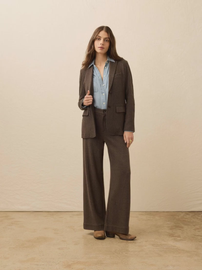 Woman is wearing a two piece brown herringbone suit with chambray shirt against a beige background