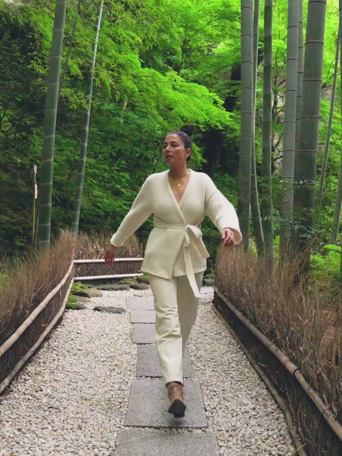 Video of a woman wearing a cream cardigan and white pants walking in a bamboo forest