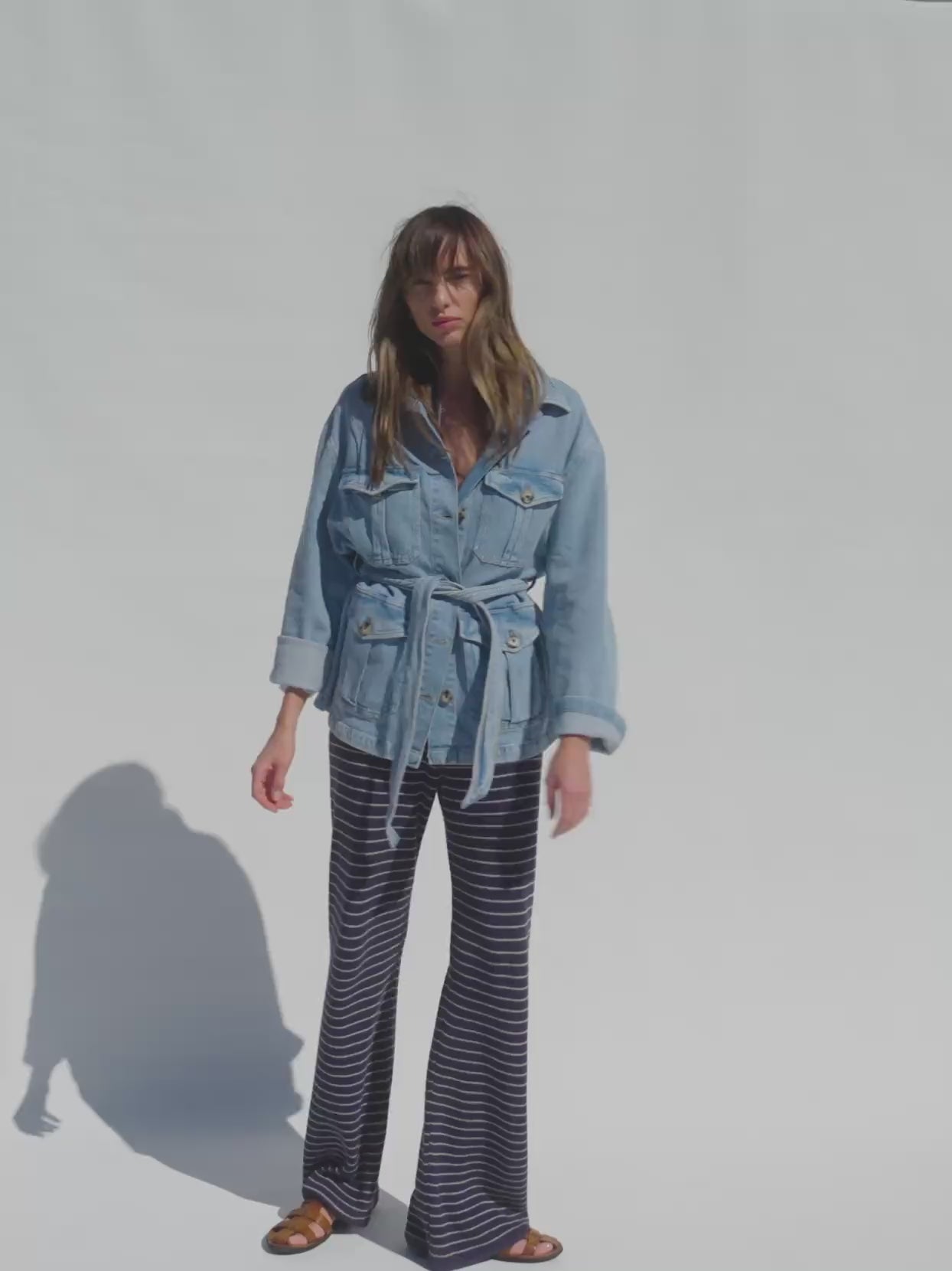 Video of a woman wearing a denim safari jacket and navy and white striped knitted pants against a white background