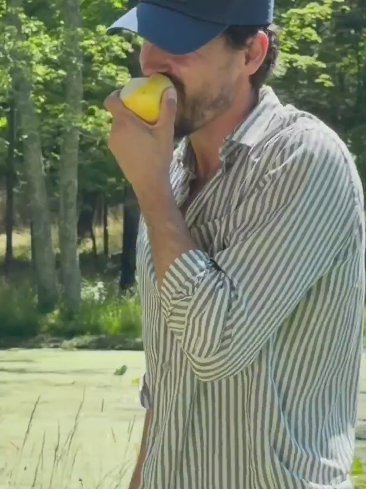 Man eating a piece of fruit wearing a striped shirt with a blue hat walking outside in nature