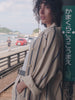 Video of a woman with curly dark hair stands on a train station platform wearing a beige tailored suit with thin dark stripes, a light gray tank top, and a black belt.