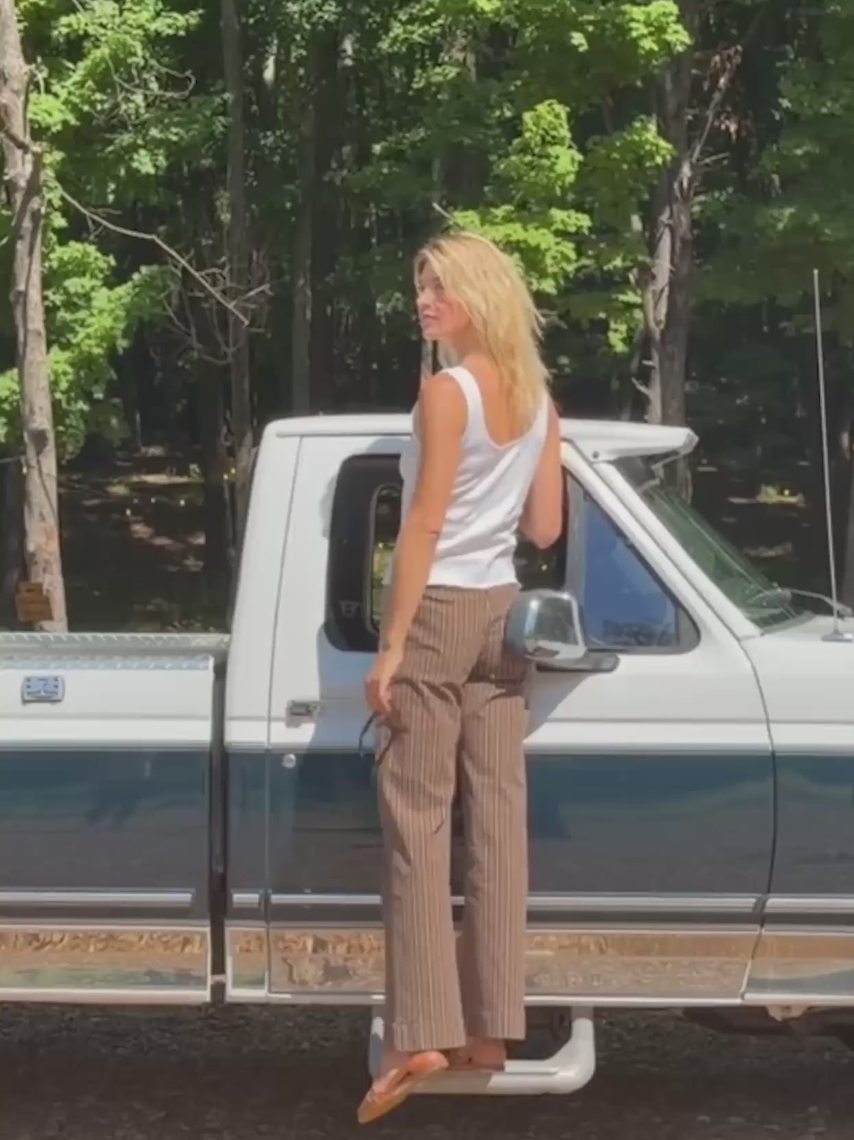 Woman wearing brown striped twill pants with a white tank top standing on a pickup truck in nature