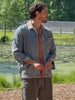 A man wearing a denim jacket over a button down shirt and brown linen shorts standing outside in nature